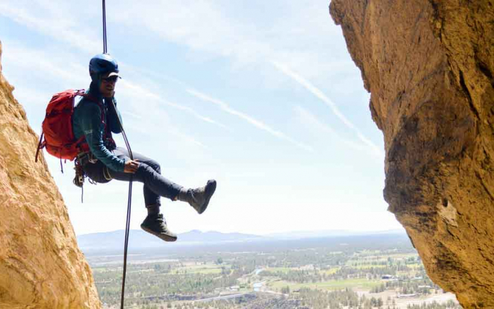 a person wearing rock climbing gear is suspended in mid-air between two rock walls. below there is a green vista with mountains in the distance. 